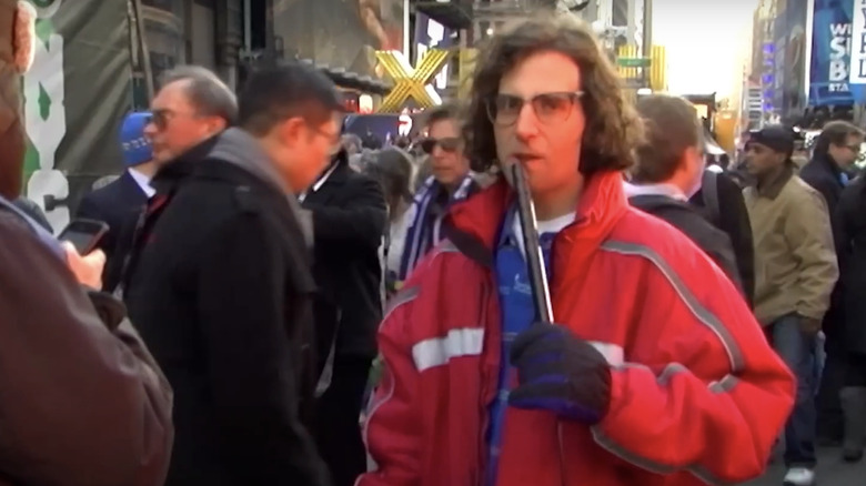 Kyle Mooney fala ao microfone enquanto está do lado de fora, vestindo um casaco vermelho, gravando uma reportagem para o Saturday Night LIve