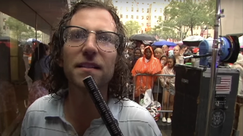 Kyle Mooney segura um microfone enquanto está do lado de fora gravando uma reportagem para o Saturday Night Live