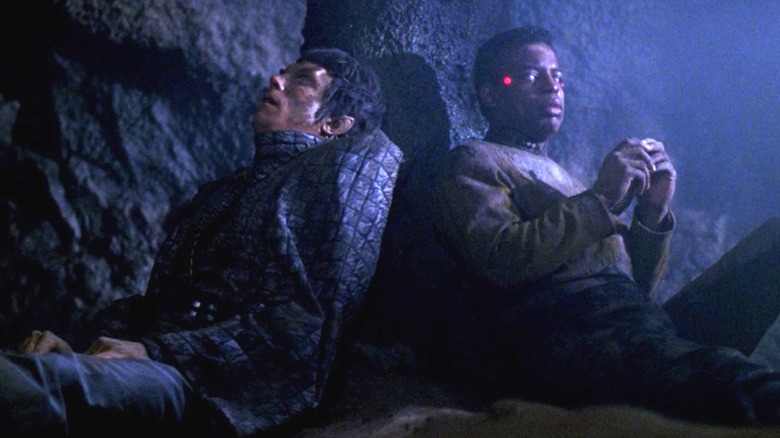 Romulano e Geordi, sentados contra um penhasco em um planeta sombrio e rochoso. O visor de Geordi está desligado. De Star Trek: a próxima geração.