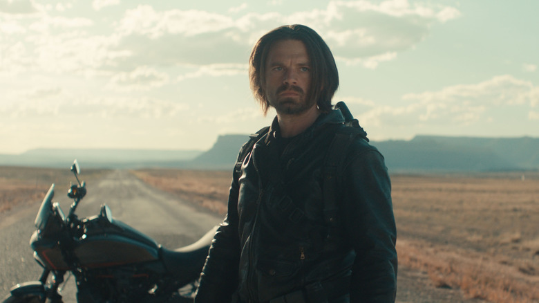 Bucky Barnes em pé em uma estrada deserta em frente a uma motocicleta estacionada