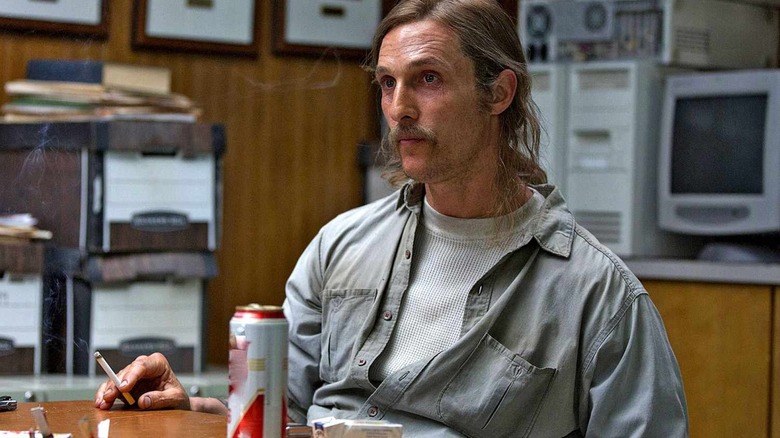 Rustin Cohle fica em uma mesa com um cigarro na mão em True Detective