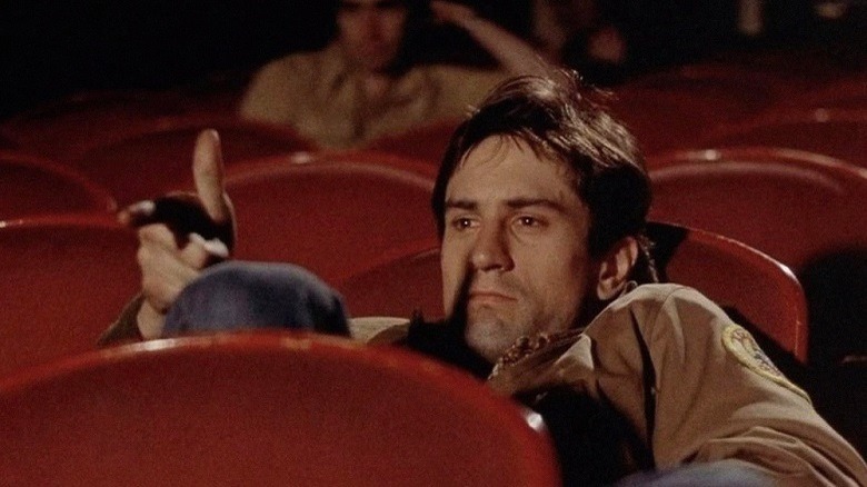 Travis Bickle sits in a half empty cinema, miming shooting at the screen