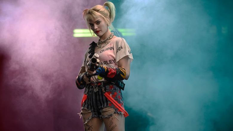 Harley Quinn stands in front of pink and blue smoke