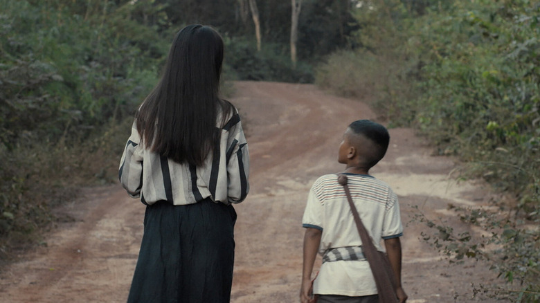 A woman walks with her son on a dirt road in The Long Walk 