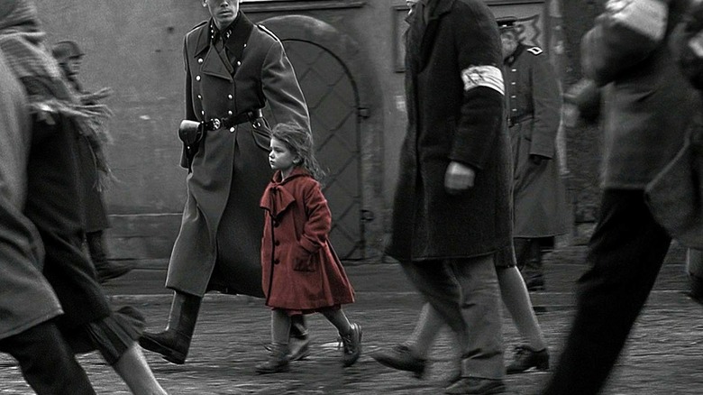 black and white image of people standing in the street with a little girl in a red coat in the foreground
