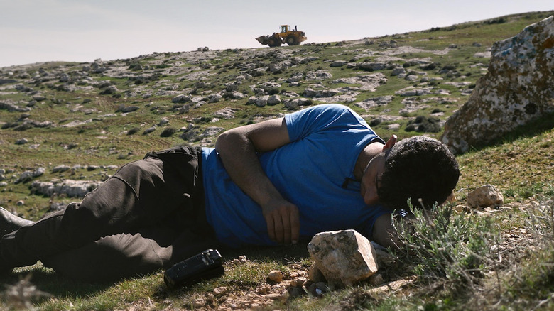 Basel Adra laying on the ground as an Israeli bulldozer demolishes a Palestinian village in the background