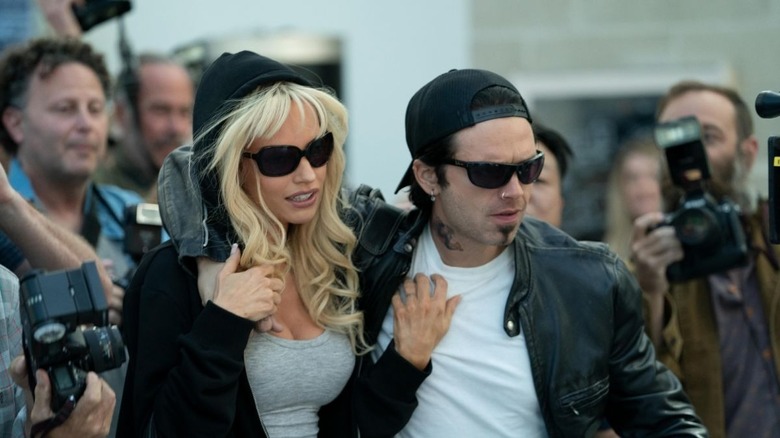 a blonde woman in sunglasses and a man in suglasses with a black cap walk through a crowd of news photographers