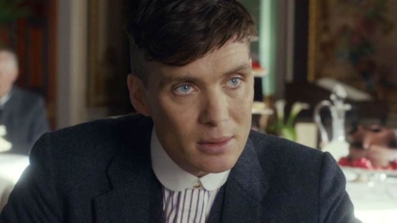 Tommy Shelby sentado em uma mesa de restaurante, olhando atentamente para alguém fora da tela em Peaky Blinders