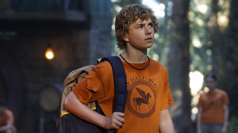 Percy stands in a forest wearing an orange Camp Half Blood shirt