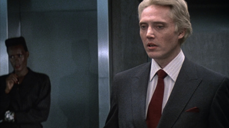 Christopher Walken's Max Zorrin stands in a suit while Grace Jones' May Day watches in the background in A View to a Kill