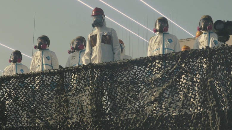 shin godzilla people wearing hazmat suits behind fence with lasers in sky