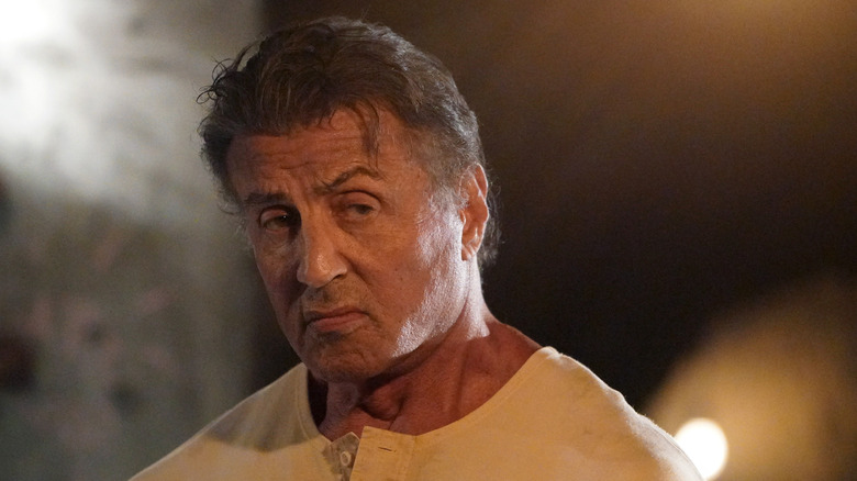 Rambo (Sylvester Stallone) looks to the right with a serious, angry expression, wearing a beige buttoned shirt, as a blurred light glows in the background in Rambo: Last Blood