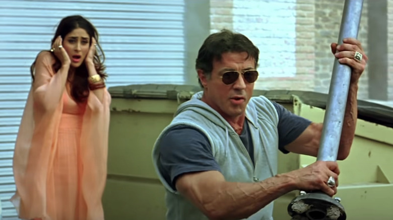 Sylvester Stallone memegang senjata sebagai Simrita Rai dari Kareena Kapoor Cowers di belakangnya di Kambakkht Ishq