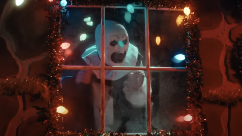 Art the clown looks excited as it looks through a window decorated with Christmas lights in terrifying 3