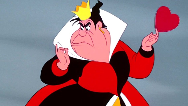 The Queen of Hearts frowns as she looks toward the left while wearing her red, white, and black gown while holding a heart outside in Alice in Wonderland