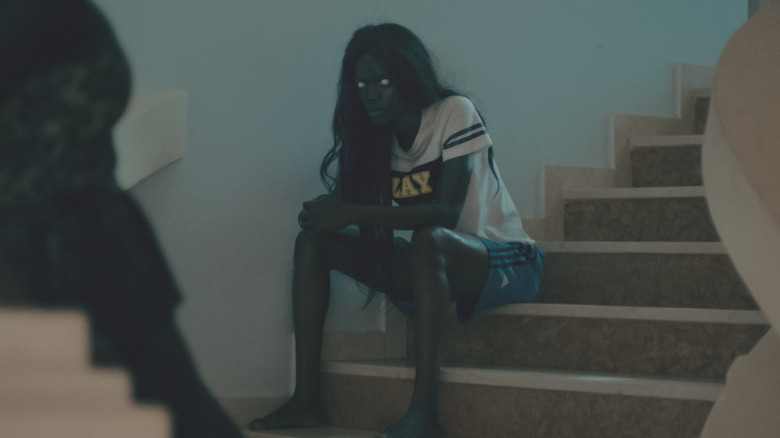 A young woman with glowing white eyes sits on steps in a sports jersey.