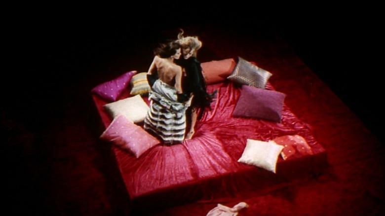 Two women caress on a red bed covered in pillows