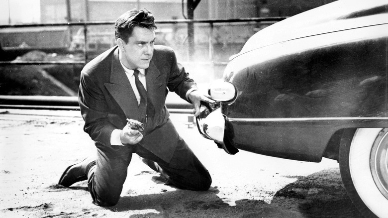 A man crouches behind a car with a gun.