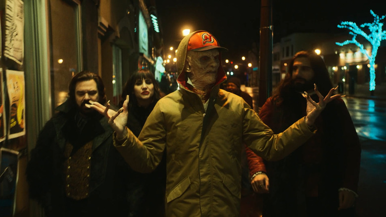 The Baron, wearing a baseball cap, walks down the street with Laszlo, Nadja, and Nandor behind him