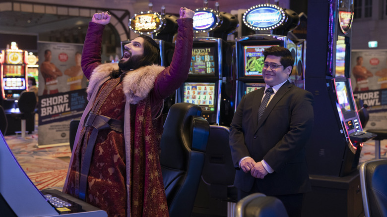 Nandor celebrates at a slot machine, Guillermo looks on bemused