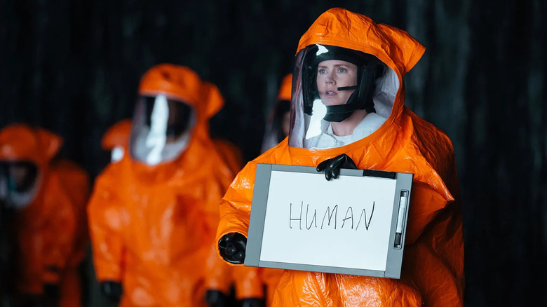 Louise Banks wears a hazmat suit and holds a sign that says "human" in a dark cave in Arrival