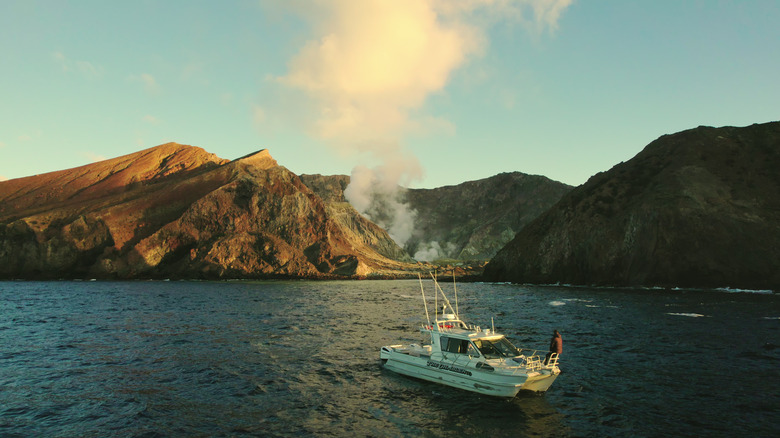 A small boat in front of a volcanic island billowing smoke