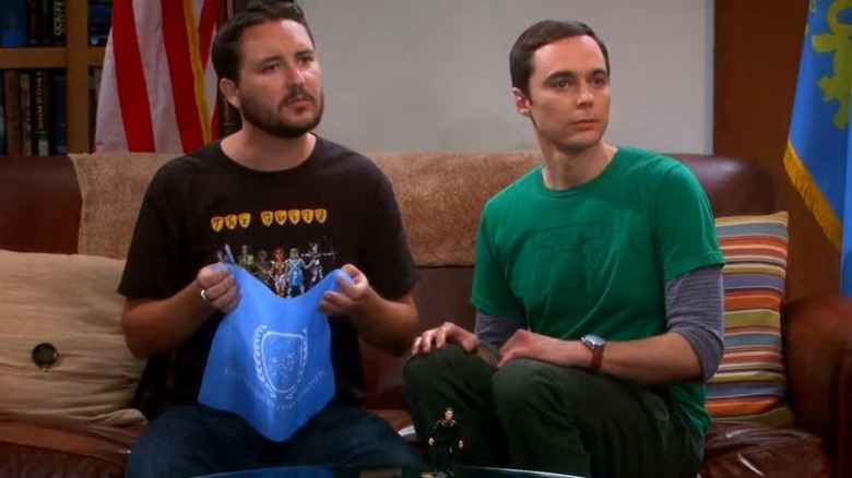 Wil Wheaton as himself holds the United Federation of Planets flag from Star Trek while sitting on a brown couch next to Jim Parsons as Sheldon Cooper on The Big Bang Theory