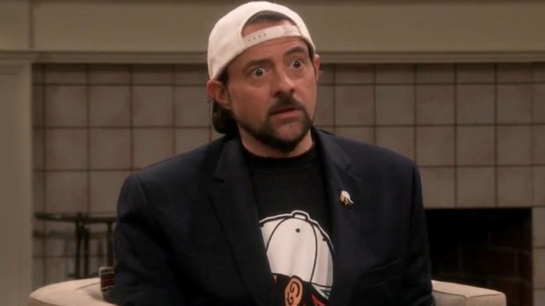 Kevin Smith as himself sits in a chair and looks ahead while wearing a white baseball hat, black blazer, and t-shirt on The Big Bang Theory