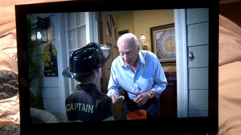 A laptop on top of a bed comforter shows a video of Buzz Aldrin as himself handing candy to a trick-or-treater dressed as a firefighter on The Big Bang Theory