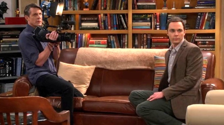 Adam Nimoy as himself holds a video camera while sitting on a brown couch with Jim Parsons as Sheldon as they both look toward the right on The Big Bang Theory