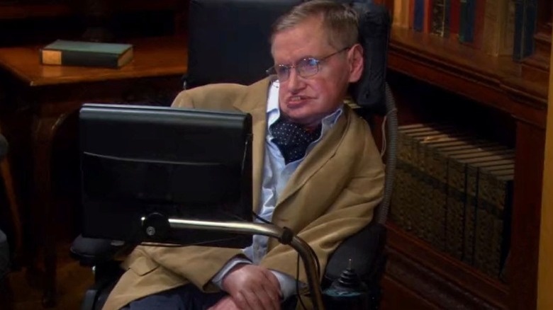 Stephen Hawking as himself sits in his wheelchair and looks ahead with bookshelves behind him on The Big Bang Theory