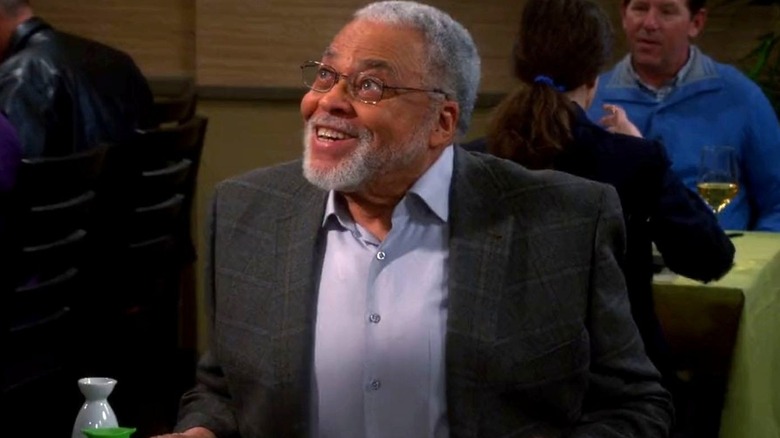 James Earl Jones as himself sits at a restaurant table and looks up smiling while wearing a gray plaid suit on The Big Bang Theory