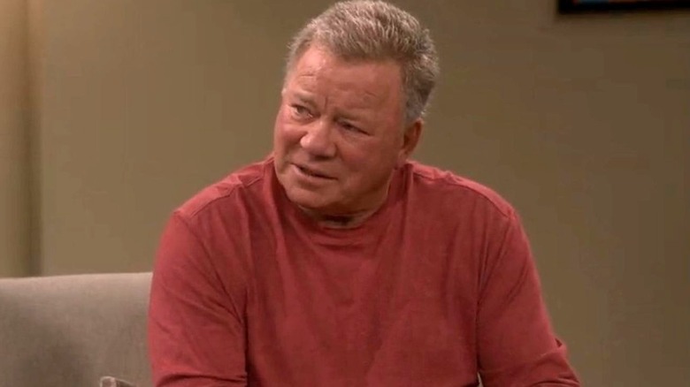 William Shatner as himself sits on a couch as he looks toward the left while wearing a red long-sleeve shirt on the Big Bang Theory