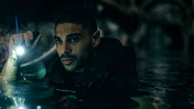 Nassim Lyes as Adil, in water up to chest height, holding a flashlight as he wades through a cave