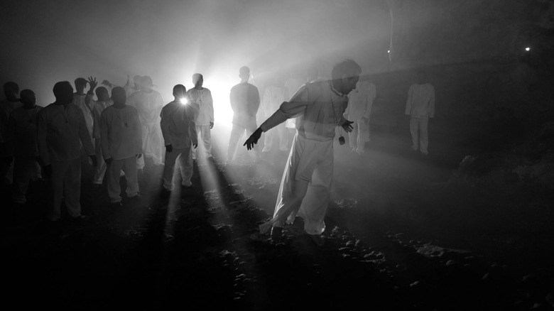 A male figure dressed in white surrounded by other men dressed in white and backlit by a spotlight in "A Lullaby to the Sorrowful Mystery"