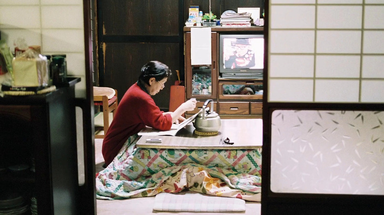 Tayoko sitting at her dining room table with a book and a pot of tea in The Works and Days (of Tayoko Shiojiri in the Shiotani Basin)