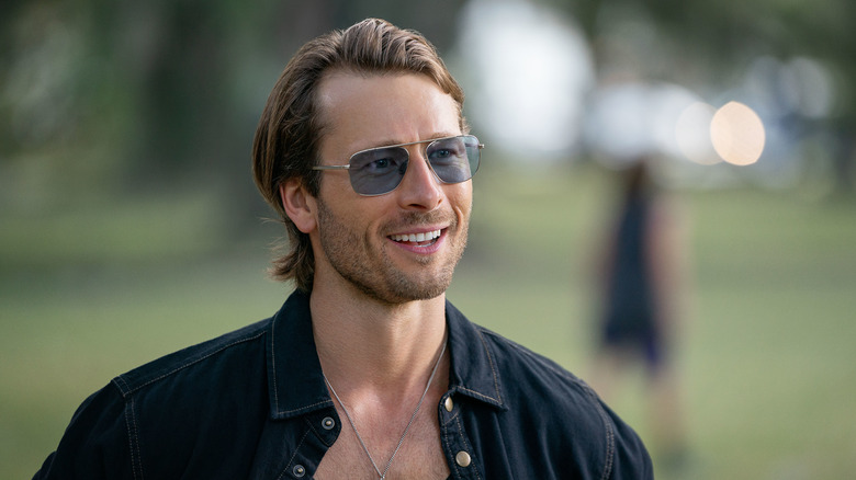 Glen Powell as Gary Johnson in Hit Man, standing in a field smiling with sunglasses on