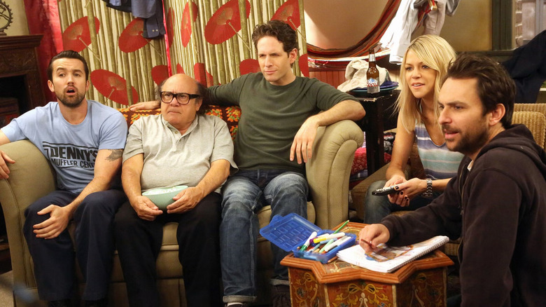 The cast of It's Always Sunny in Philadelphia sitting in a living room, looking at a TV