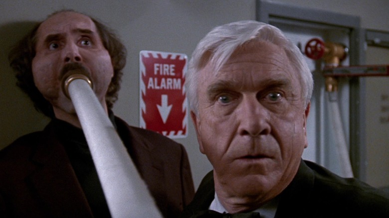 Leslie Nielsen as Frank Drebin is panicked as a man is inflated with a fire hose behind him in The Naked Gun