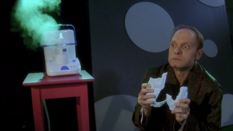 Niles kneels next to a bottle warmer, looking worried after breaking a porcelain baby during a dream sequence