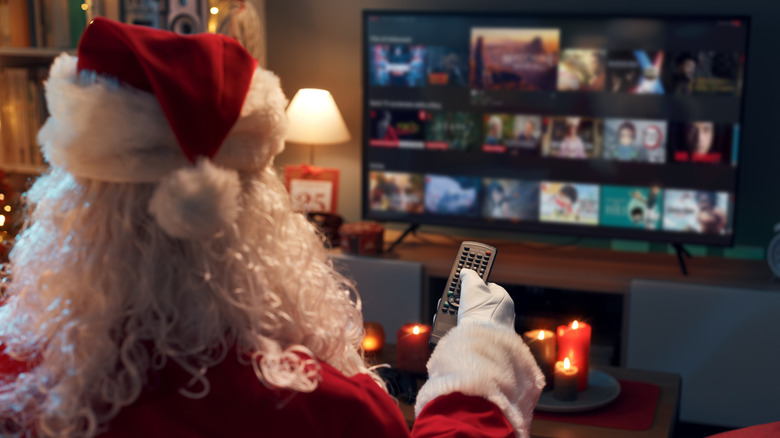 A stock photo of Santa Claus sitting on his living room couch, holding a TV remote and selecting a movie to watch