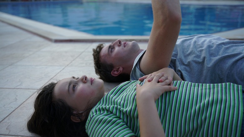 Sophie and Calum lie on the concrete patio next to a pool, Calum's arm raised and Sophie's hands clasped across her chest