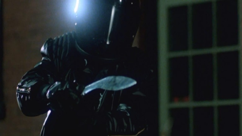 A person wearing black leather and a motorcycle helmet points a large knife at the camera