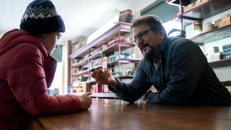 Tom Savini on Locke & Key behind a counter and giving an item to a child.