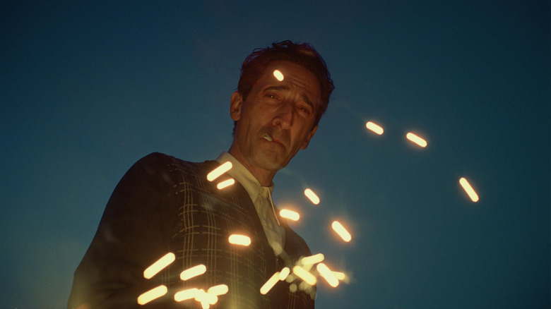 Adrien Brody as László Tóth smoking a cigarette in front of fiery sparks in a scene from The Brutalist