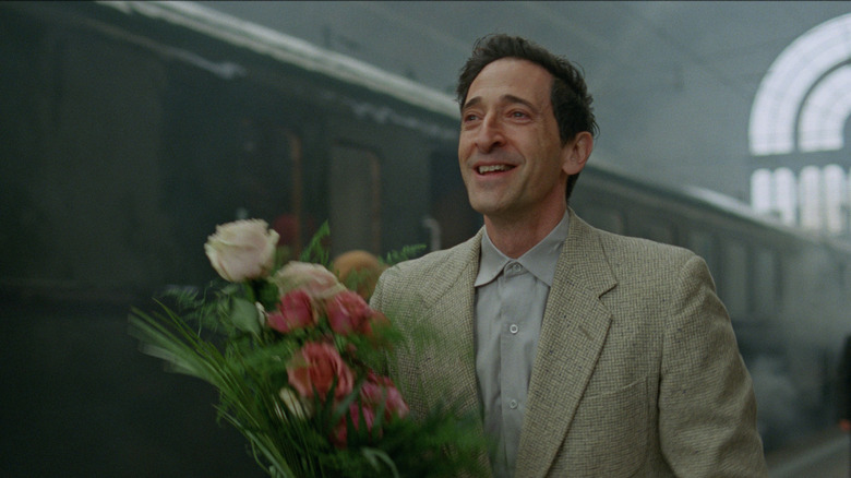 Adrien Brody as László holds flowers at a train station in The Brutalist