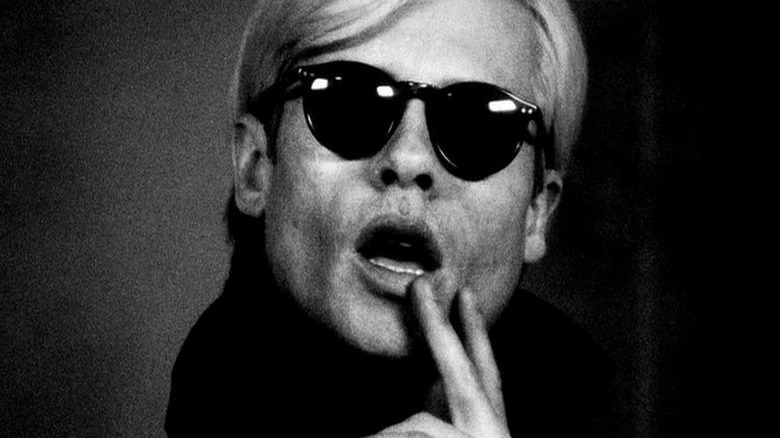 close up of man with white hair and sunglasses with his mouth open holding two fingers to his face