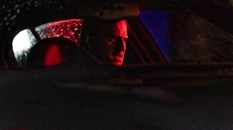 Oz Cobb's face is highlighted in dramatic red light as he sits in the car