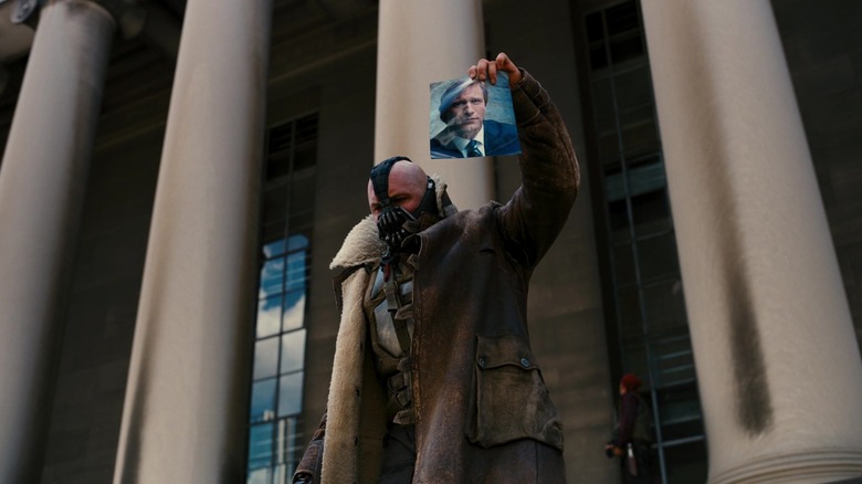 Tom Hardy as Bane wearing a leather mask and jacket while holding a picture of Harvey Dent from The Dark Knight Rises