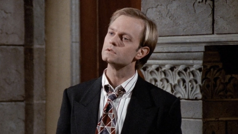 David Hyde Pierce's Niles stand in front of a stone fireplace in a suit in Frasier